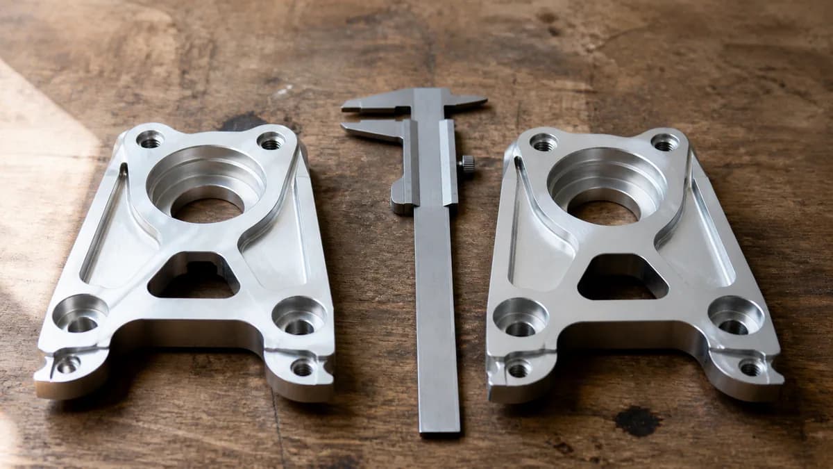 Editorial photograph of two near-identical machined metal parts laid side by side on a workbench with a pair of calipers between them